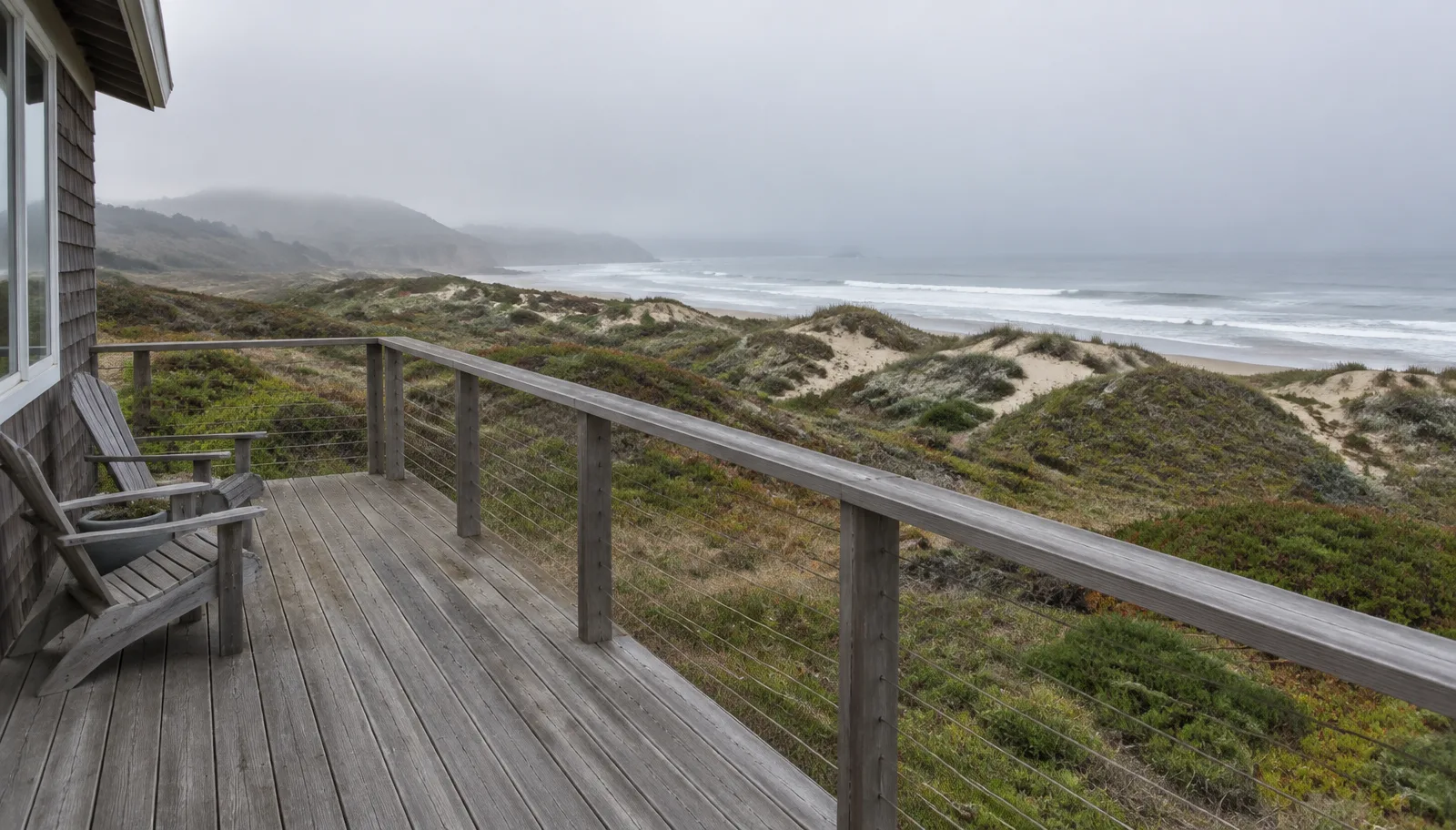 A weathered wood deck with simple railings overlooking coastal dunes and a foggy Pacific Ocean
