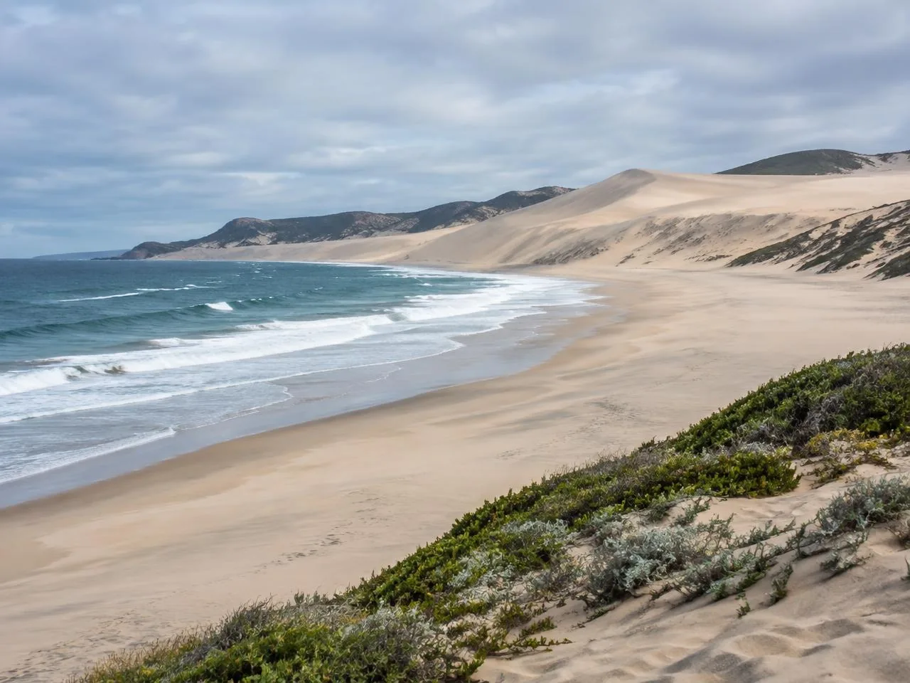 A wide, uncrowded beach backed by tall sand dunes under a soft overcast sky